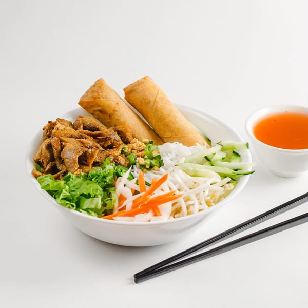 A Vietnamese vermicelli bowl with grilled pork, egg rolls, and fresh vegetables, with a side of sauce.