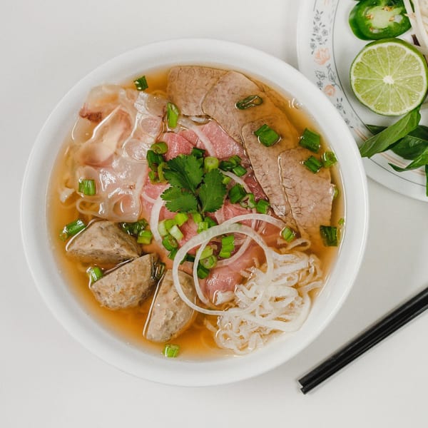 A bowl of Vietnamese pho with beef, meatballs, noodles, and fresh herbs, with lime and jalapeños on the side.